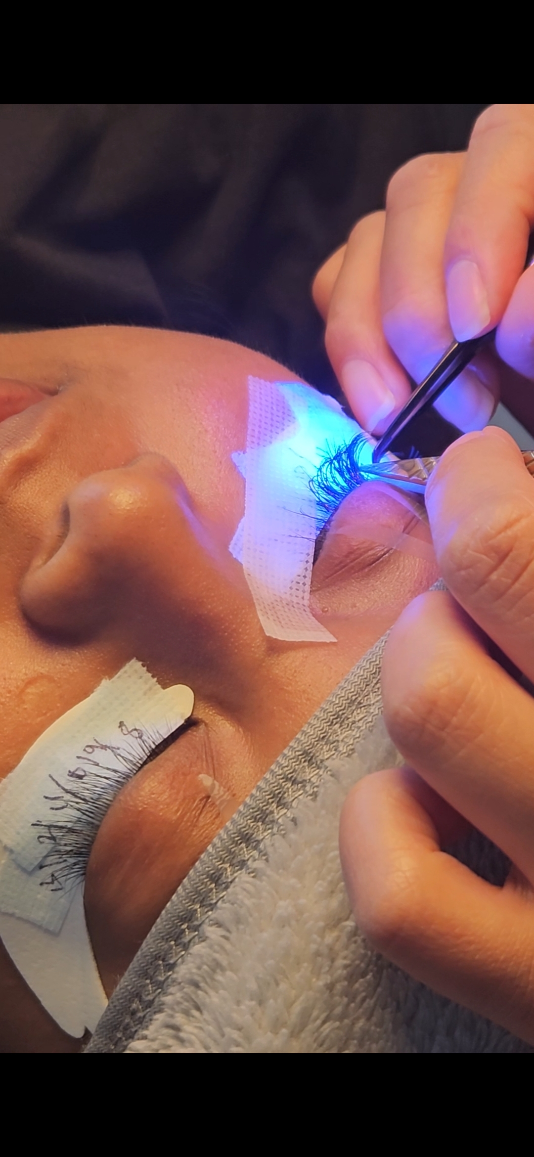 Lash artist applying extensions with precision tweezers under a curing light